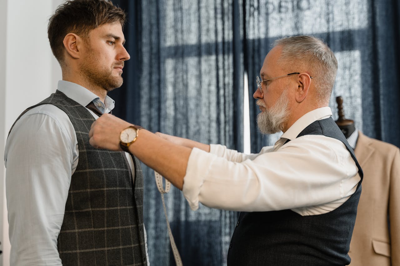 hero-img-02 Professional tailor measuring a client for a custom suit in a workshop setting.