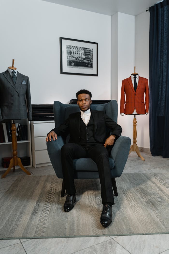 gallery-1 A fashionable man seated in a chair, surrounded by couture suits, exuding style and confidence.